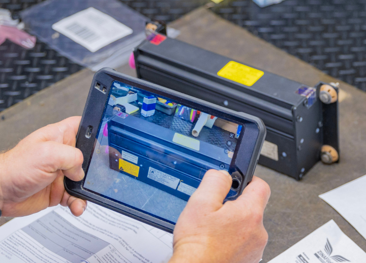 A person photographing a black electronic device on a table with a tablet.