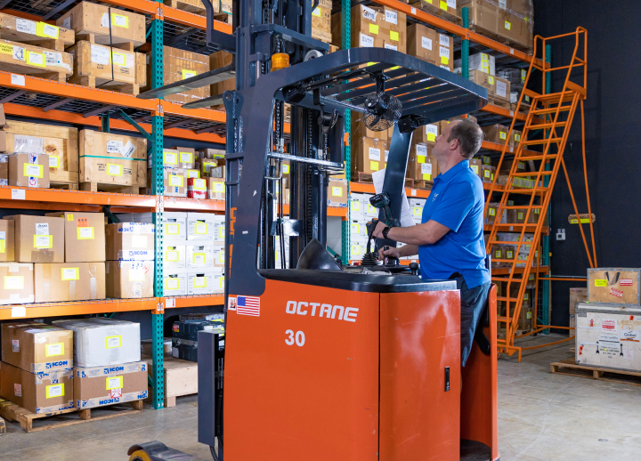 A person operating an orange forklift in a warehouse with shelves full of boxes.