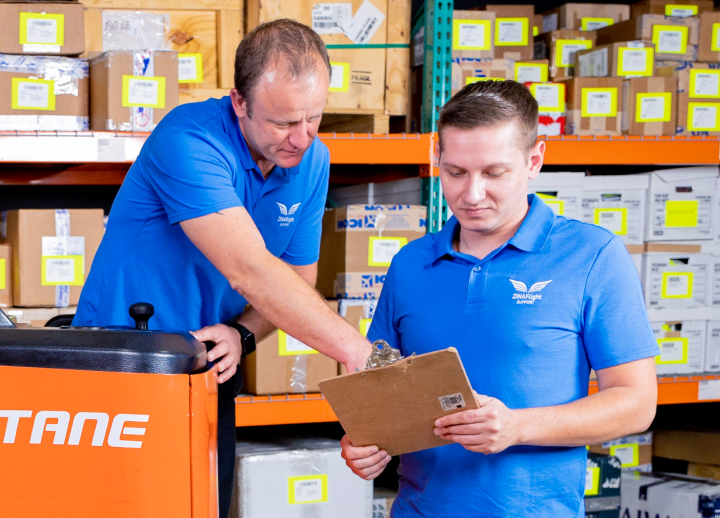 Two warehouse workers in blue Zina Flight shirts reviewing a clipboard in a storage area with shelves filled with boxes.