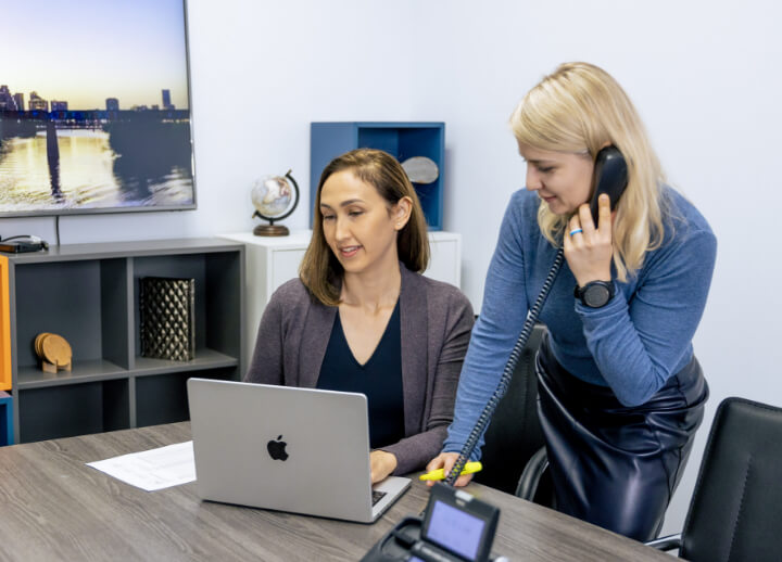 Two women in an office, one using a laptop and the other standing and talking on the phone.